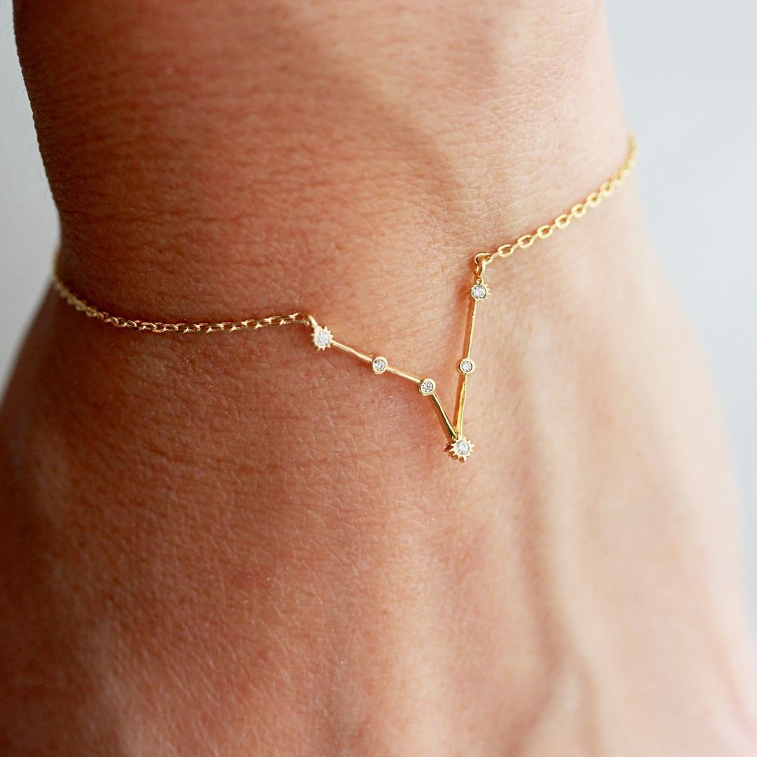 Een close-up van een delicate gouden AstroChic Sterrenarmband, ontworpen in de vorm van een sterrenbeeld met sprankelende kristallen. De armband straalt verfijning en elegantie uit en is verstelbaar voor een perfecte pasvorm. Ideaal als persoonlijk en stijlvol accessoire voor astrologieliefhebbers.