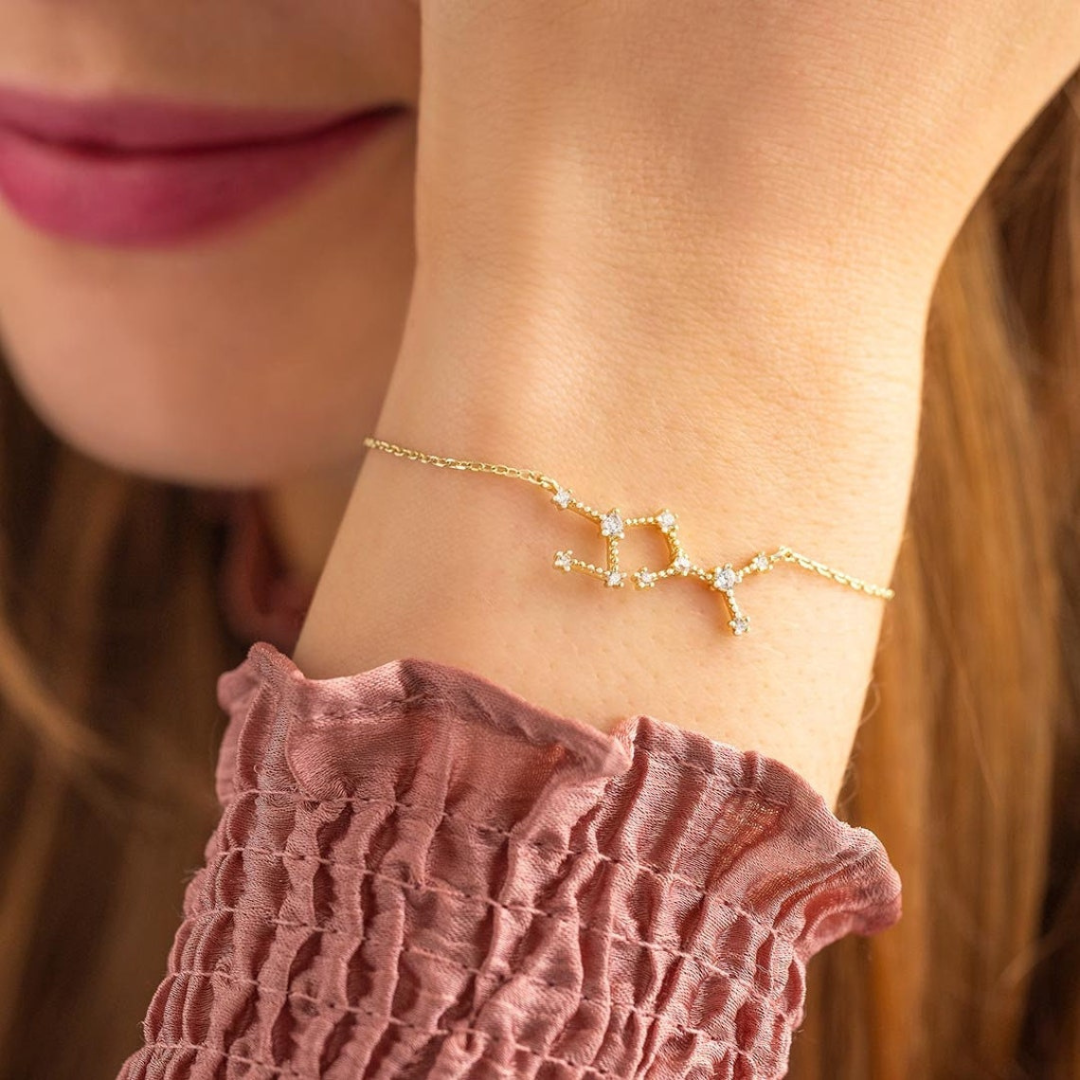 Een close-up van een delicate gouden AstroChic Sterrenarmband, geïnspireerd door sterrenbeelden en bezet met glinsterende kristallen. De armband, gedragen om de pols van een vrouw in een elegante roze blouse, straalt verfijning en persoonlijkheid uit. Perfect als uniek en verstelbaar sieraad voor liefhebbers van astrologie en tijdloze stijl.