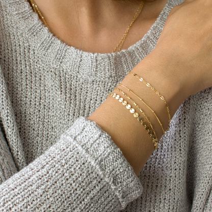 Een stijlvolle close-up van een vrouw die meerdere fijne gouden Aurora & Blossom armbanden draagt, met delicate details en een elegante uitstraling. Deze minimalistische armbanden stralen tijdloze charme uit en zijn perfect om elke outfit te verfraaien.