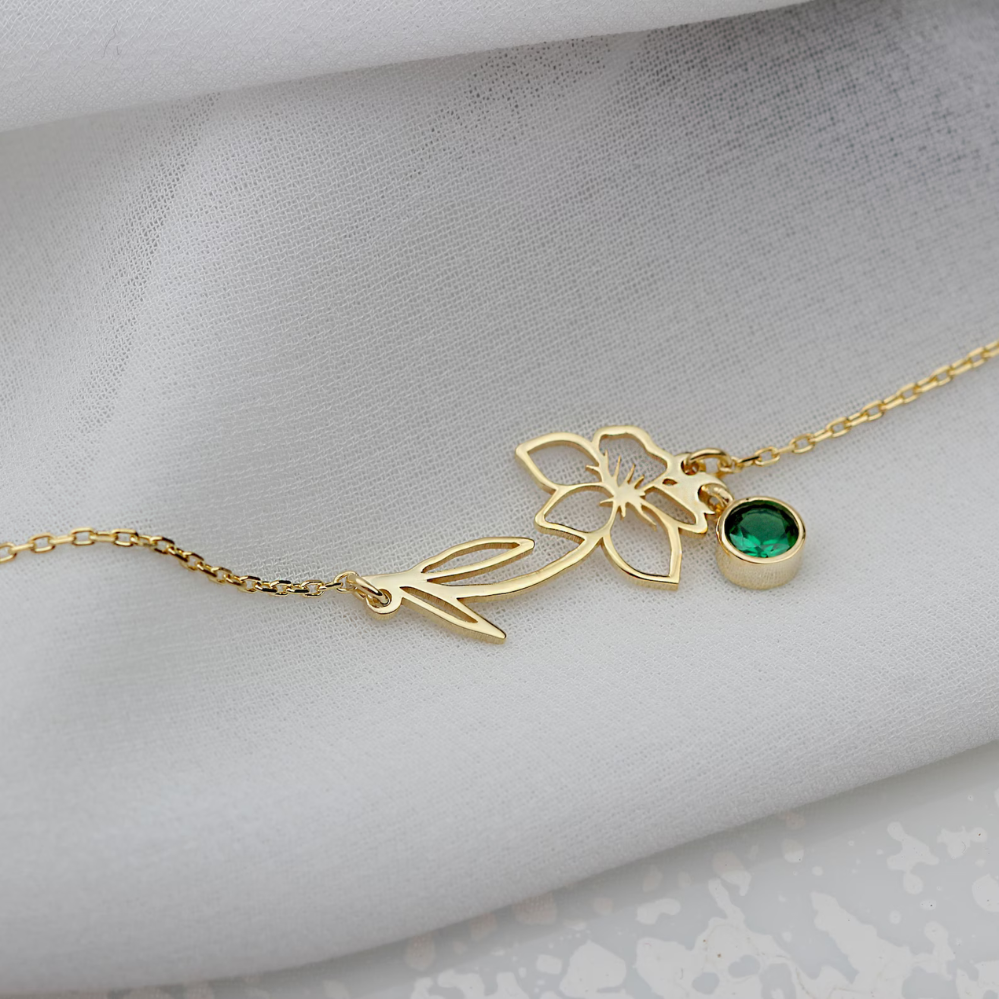 Een verfijnde gouden Blooming Essence armband met een delicate bloemvorm en een groene edelsteenhanger, subtiel gepresenteerd op een zachte witte achtergrond. Deze persoonlijke en elegante armband is een perfecte keuze voor een stijlvolle en unieke look.