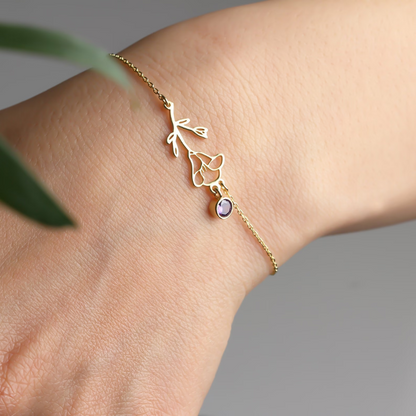 Een elegante gouden Blooming Essence armband, subtiel gedragen om de pols, met een sierlijke bloemvorm en een paarse edelsteenhanger. Deze persoonlijke en verfijnde armband straalt stijl en uniekheid uit, ideaal voor elke gelegenheid.