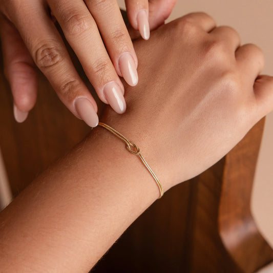 Een close-up van een delicate gouden EleganceVibes armband, met een verfijnde knoopdetail, subtiel gedragen om de pols van een vrouw met perfect verzorgde nagels. Deze tijdloze en stijlvolle armband straalt elegantie uit en is ideaal voor zowel dagelijks gebruik als speciale gelegenheden.