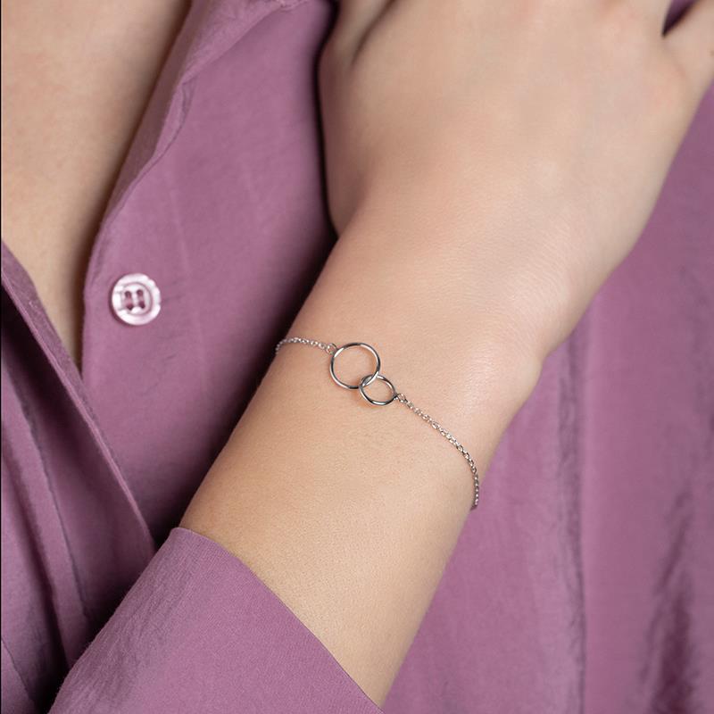 Een elegante close-up van een zilveren Eternale Unité armband, met twee in elkaar grijpende ringen als symbool van verbondenheid en tijdloosheid. Gedragen om de pols van een vrouw in een stijlvolle paarse blouse, straalt deze verfijnde armband een subtiele en moderne elegantie uit. Perfect voor elke gelegenheid.