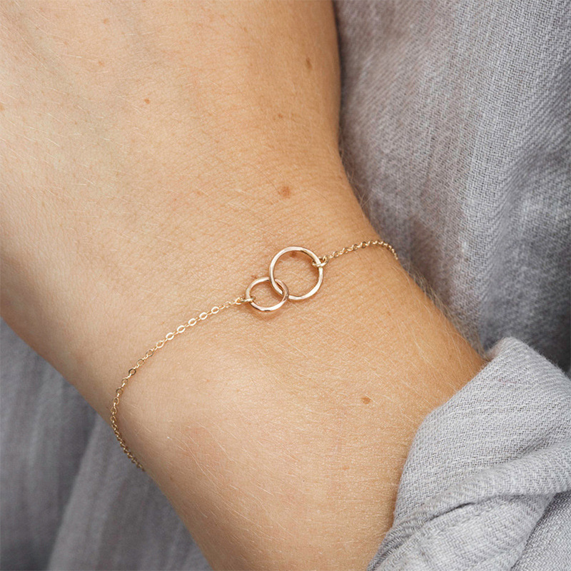 Een close-up van de gouden Eternale Unité armband, met twee verfijnde in elkaar grijpende ringen als symbool van verbondenheid en tijdloze elegantie. Deze minimalistische armband straalt subtiliteit en verfijning uit, perfect voor dagelijks gebruik of speciale gelegenheden.