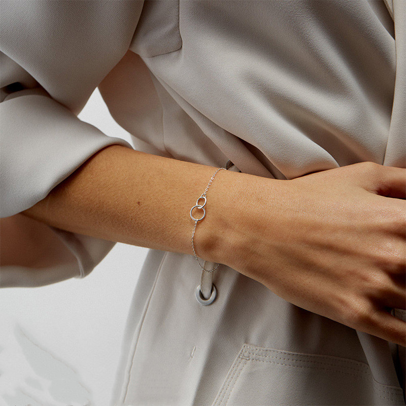 Een stijlvolle zilveren Eternale Unité armband met twee in elkaar grijpende ringen, gedragen door een vrouw in een elegante, lichtgrijze outfit. Deze tijdloze en minimalistische armband straalt verfijning en verbondenheid uit, ideaal voor zowel dagelijks gebruik als speciale gelegenheden.