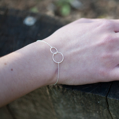 Een minimalistische zilveren Eternale Unité armband met twee in elkaar grijpende ringen, subtiel gedragen om de pols van een vrouw. Deze tijdloze en elegante armband straalt verfijning uit en is perfect als symbool van verbondenheid voor elke gelegenheid