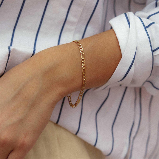 Een stijlvolle gouden Lusso Vero armband met een verfijnd kettingontwerp, gedragen om de pols van een persoon in een wit met blauw gestreepte blouse. Perfect om een tijdloze elegantie aan elke outfit toe te voegen.