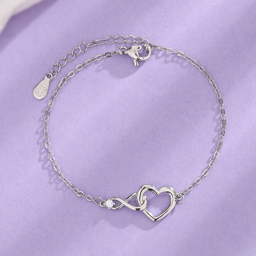 Een zilverkleurige Lustre Luxe armband met een elegant hart-en-infinity-ontwerp, voorzien van een subtiele glinsterende steen, gepresenteerd op een zachte paarse achtergrond. Perfect voor een tijdloze en verfijnde stijl.