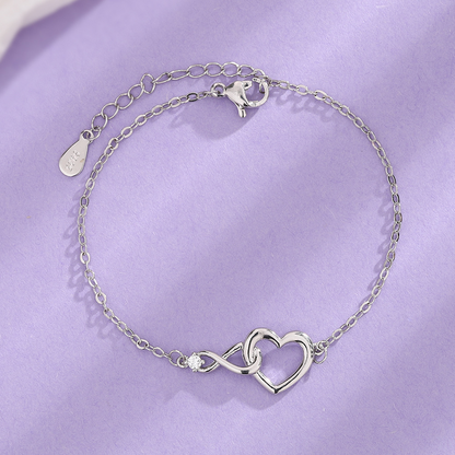 Een zilverkleurige Lustre Luxe armband met een elegant hart-en-infinity-ontwerp, voorzien van een subtiele glinsterende steen, gepresenteerd op een zachte paarse achtergrond. Perfect voor een tijdloze en verfijnde stijl.