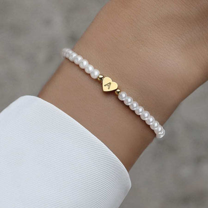 Een close-up van een verfijnde parelarmband met een gouden hartvormige hanger waarin de letter "A" is gegraveerd. De Double Love Heart Bracelet combineert elegantie met een persoonlijke touch, perfect voor een tijdloze en stijlvolle uitstraling bij elke gelegenheid.